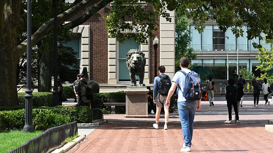 Several university students walk on campus with Scholars' Lions sculpture in the background at Columbia University. 