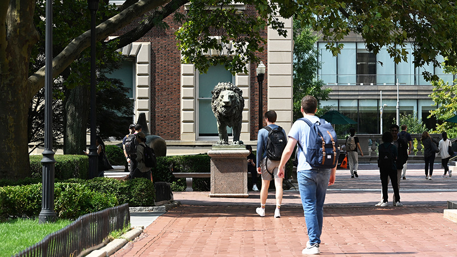Several university students walk on campus with Scholars' Lions sculpture in the background at Columbia University. 