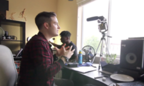 Man seated at a desk in front of a window recording video of himself. Another man in the background